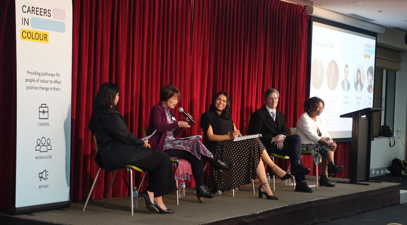 A Careers in Colour panel event featuring five speakers seated on stage against a red curtain backdrop, discussing racial diversity in the public sector, with a banner displaying the organisation’s mission to create pathways for people of colour.
